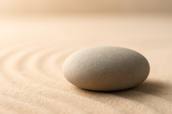Ein glatter, runder Stein ruht auf fein geharktem, beigem Sand mit sanften Wellenmustern – Zen-Garten-Atmosphäre.