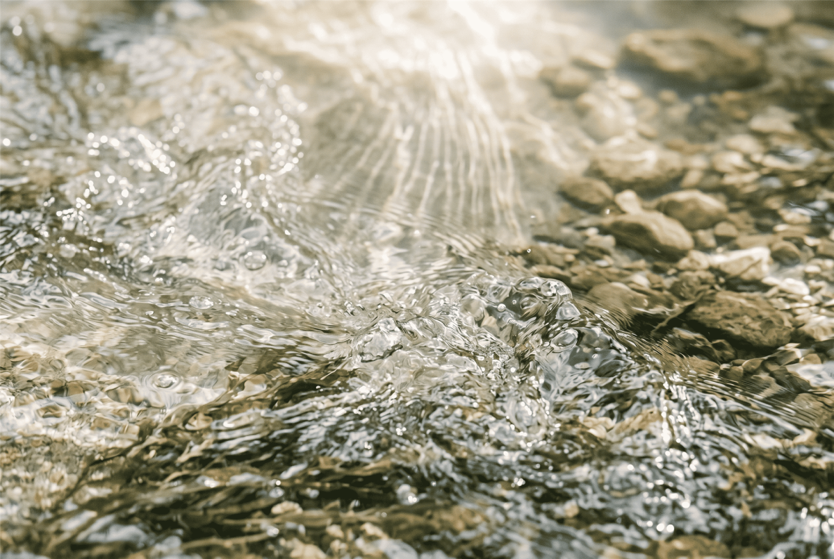 Nahaufnahme von aufgewirbeltem, klarem Wasser mit brechenden Lichtstrahlen über Kieselsteinen zur Visualisierung von Entlastung beim Breathwork.